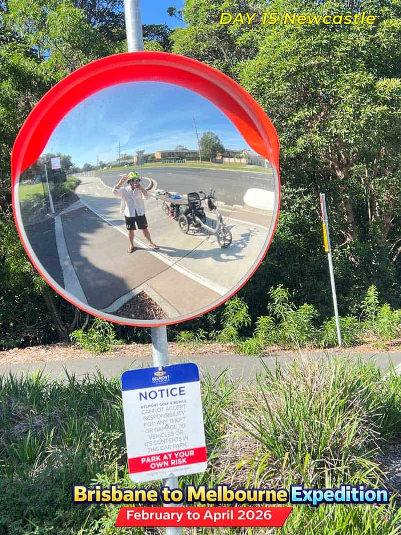 A reflection of a person riding an adult tricycle on a road. The tricycle and rider are visible in a convex mirror.