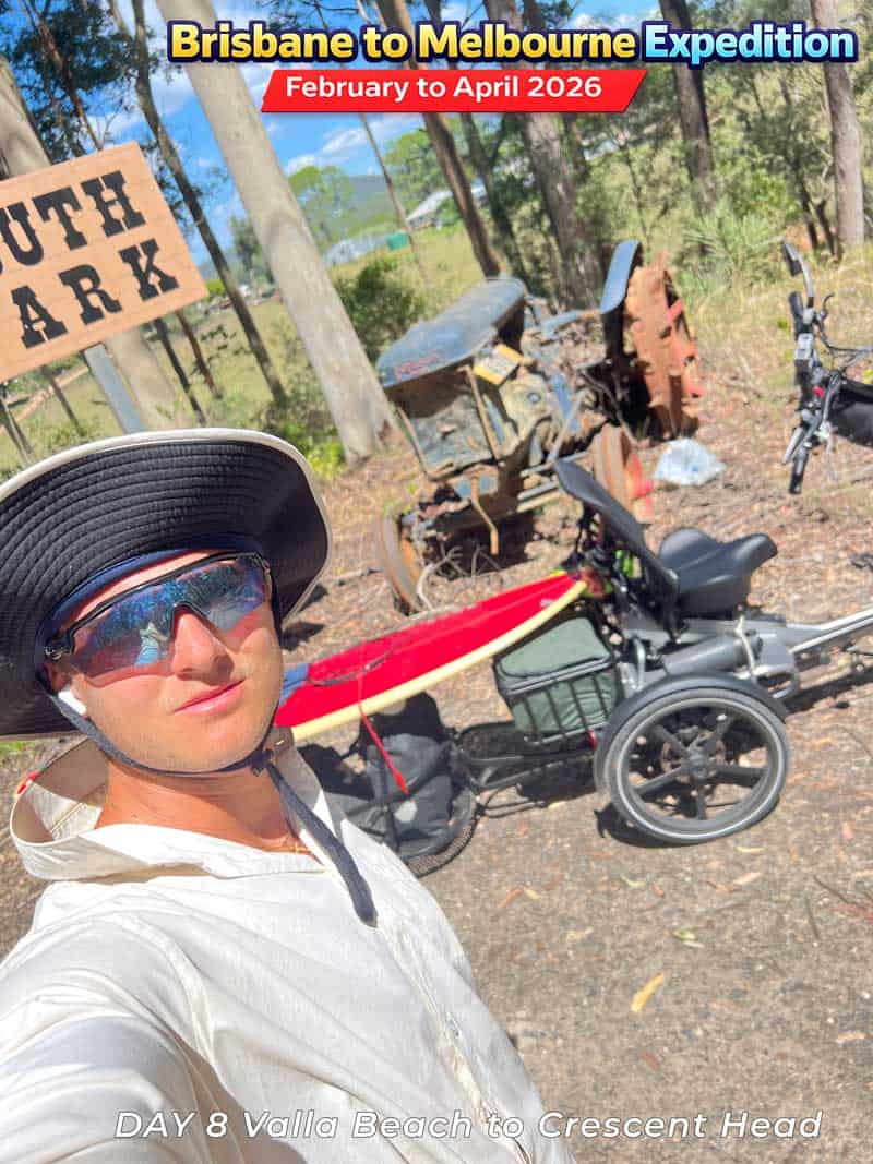 Man taking selfie with adult tricycle and surfboard. The tricycle has a large basket and is parked on dirt path.
