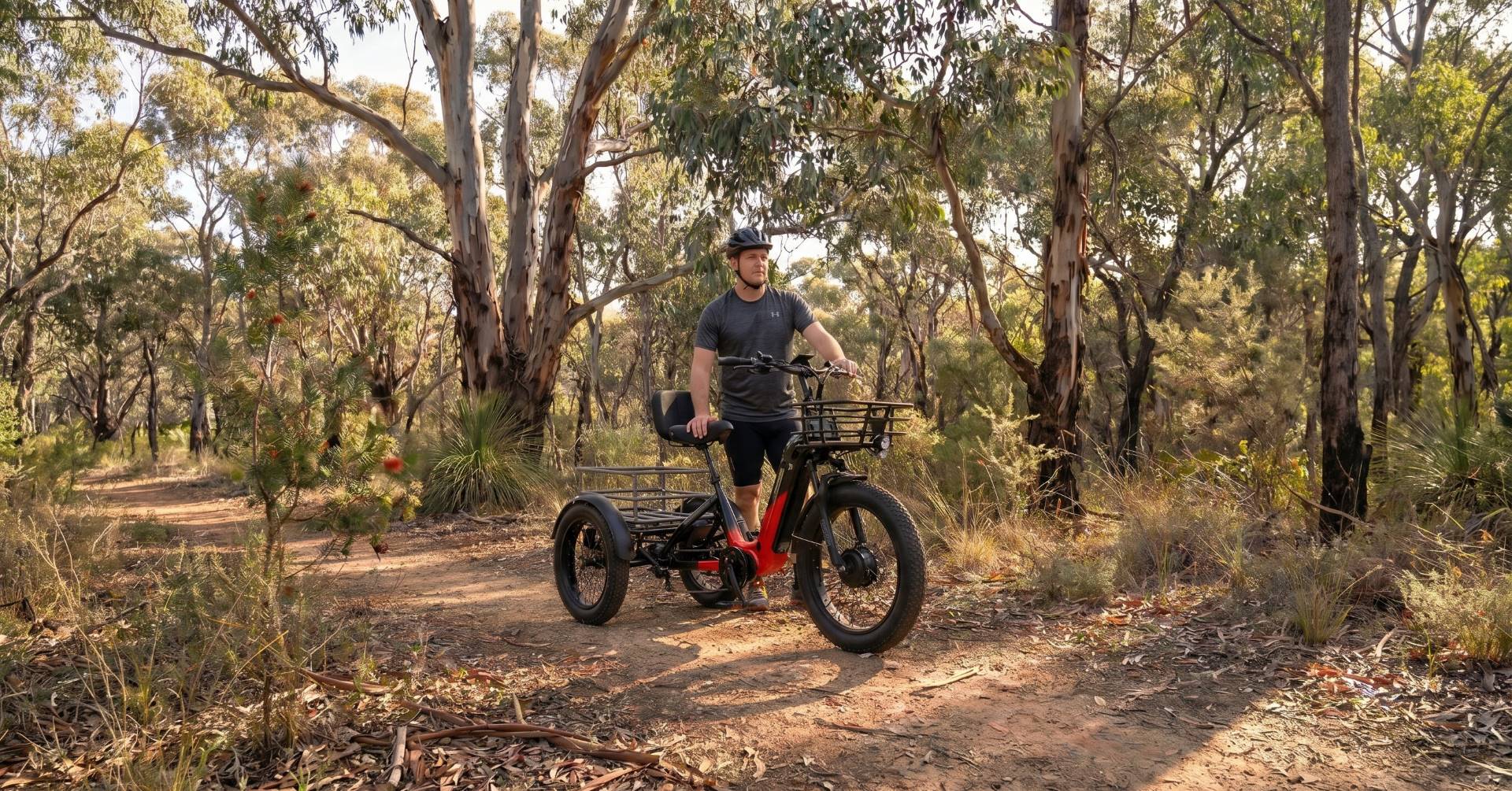 A man rides an orange electric adult tricycle on a dirt track in the Australian bush. The trike has three wheels and a sturdy frame.