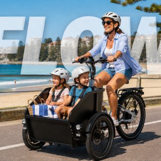A woman pedalling a black cargo tricycle along a coastal path, transporting two children securely in the front box.